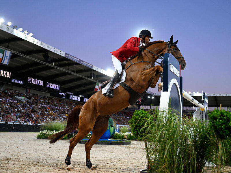 Rider in red and white attire on a horse, jumping over an obstacle in an equestrian event with spectators in the background.