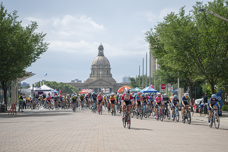 A group of cyclists participating in an event with tents and flags on the side, and a large domed building in the background.