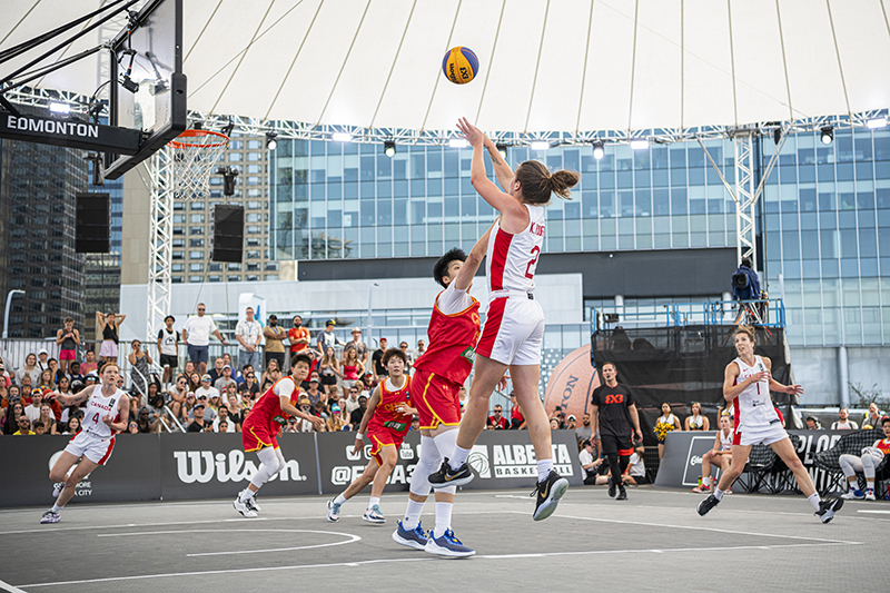 A basketball player jumping to reach the ball during an outdoor game and a crowd of spectators in the background.