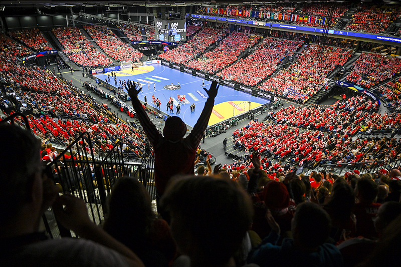 An enthusiastic spectator at a large indoor handball event, captured from behind with arms raised in a gesture of excitement.