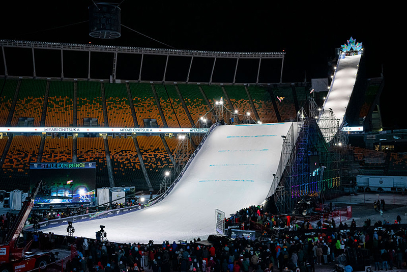 A night scene at a large outdoor stadium, featuring an illuminated ramp for snow sports events, surrounded by spectators.